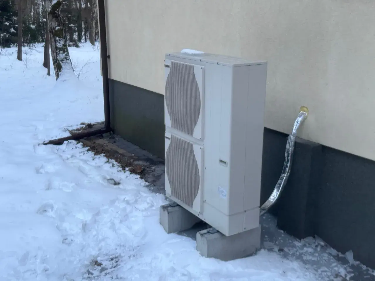Jednostka zewnętrzna pompy ciepła stoi na betonowych podstawach w zasypanym śniegiem ogrodzie. Czy pompa ciepła jest głośna? Zimą, gdy wszystko wokół jest ciche, można to sprawdzić.