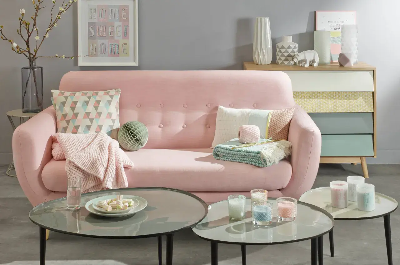 Sofá rosa pastel con cojines y mantas, mesas de centro con velas y malvaviscos, y un mueble de cajones de colores.
