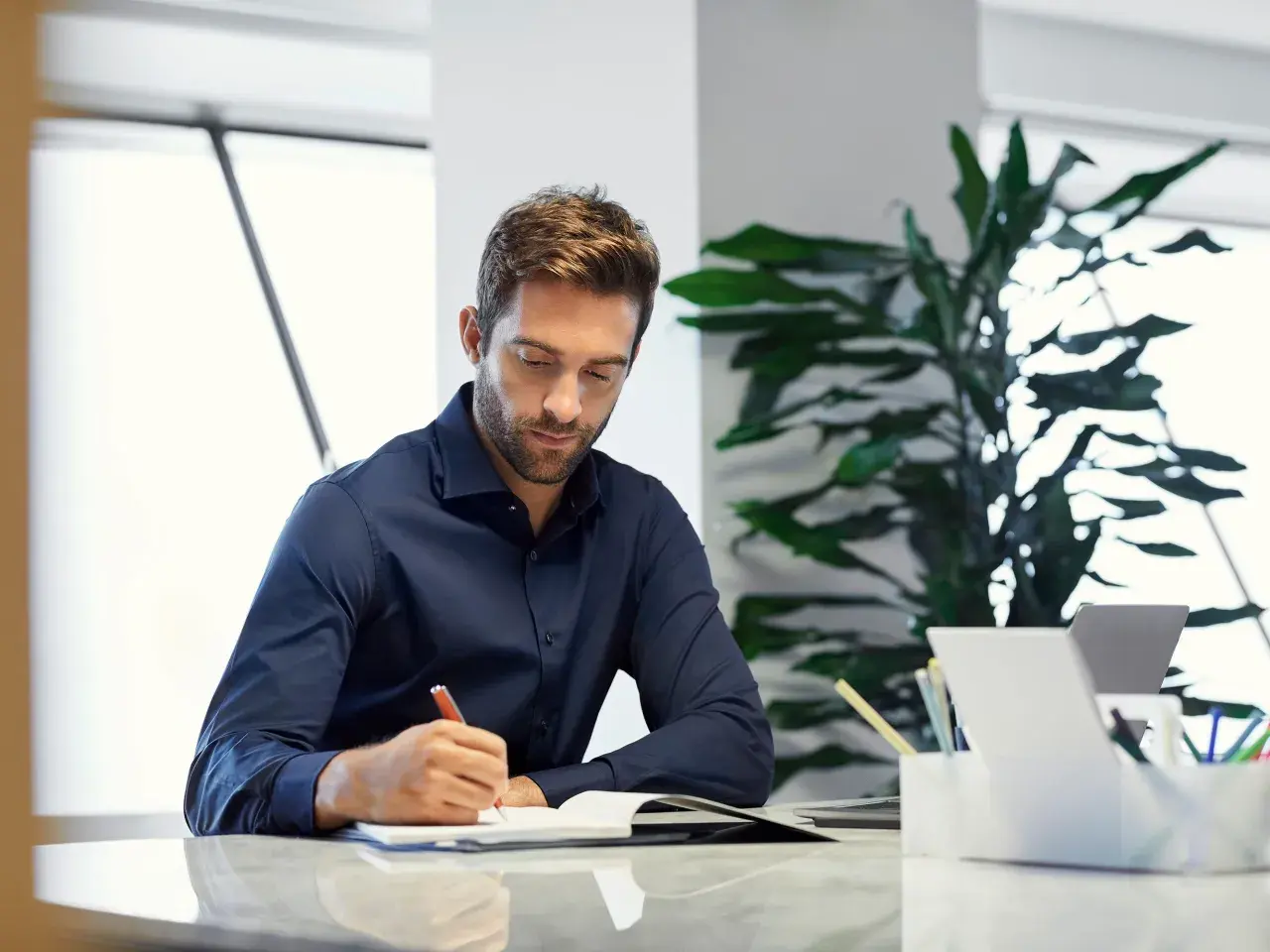 Hombre concentrado escribiendo en un diario en su escritorio, rodeado de plantas y un portátil.