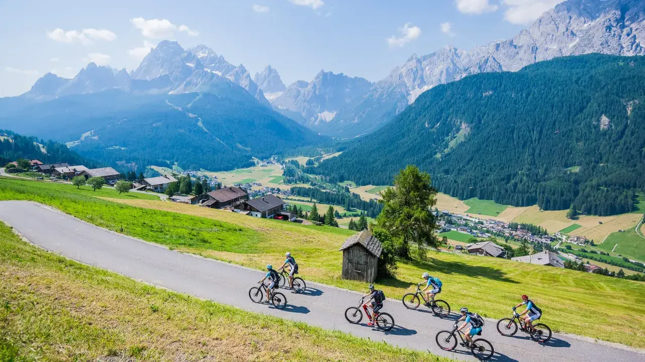 A group enjoys a cycling holiday in the Dolomites, riding down a scenic road with stunning mountain views and a charming village below.