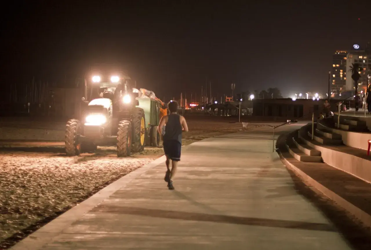 Mężczyzna uprawia bieganie w nocy na promenadzie, obok pracującego traktora na plaży.