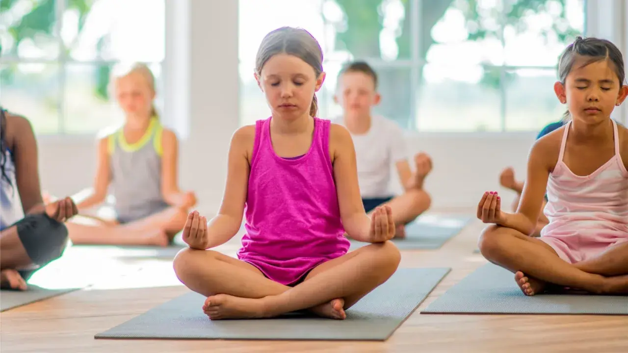 Niños practicando ejercicios de respiración y meditación en un salón de clases, sentados en colchonetas con los ojos cerrados.