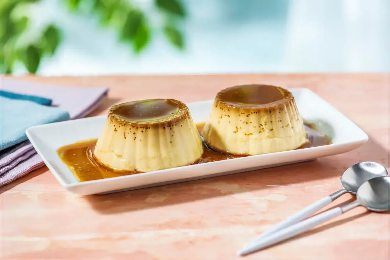 Zwei köstliche flan caramel Desserts auf einem weißen Teller, mit flüssiger Karamellsauce übergossen. Löffel liegen daneben.