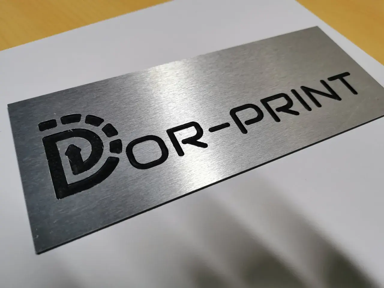 Metalowa tabliczka grawer z logo "Dor-Print". Wygląda jakby była wykonana z aluminium.
