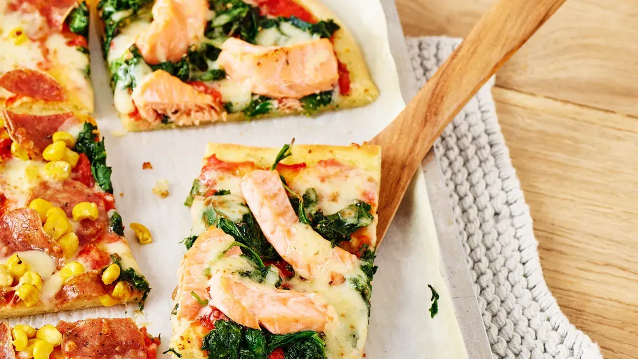 Pizza mit Lachs und Spinat, belegt mit geschmolzenem Käse, wird mit einem Holzschieber serviert.