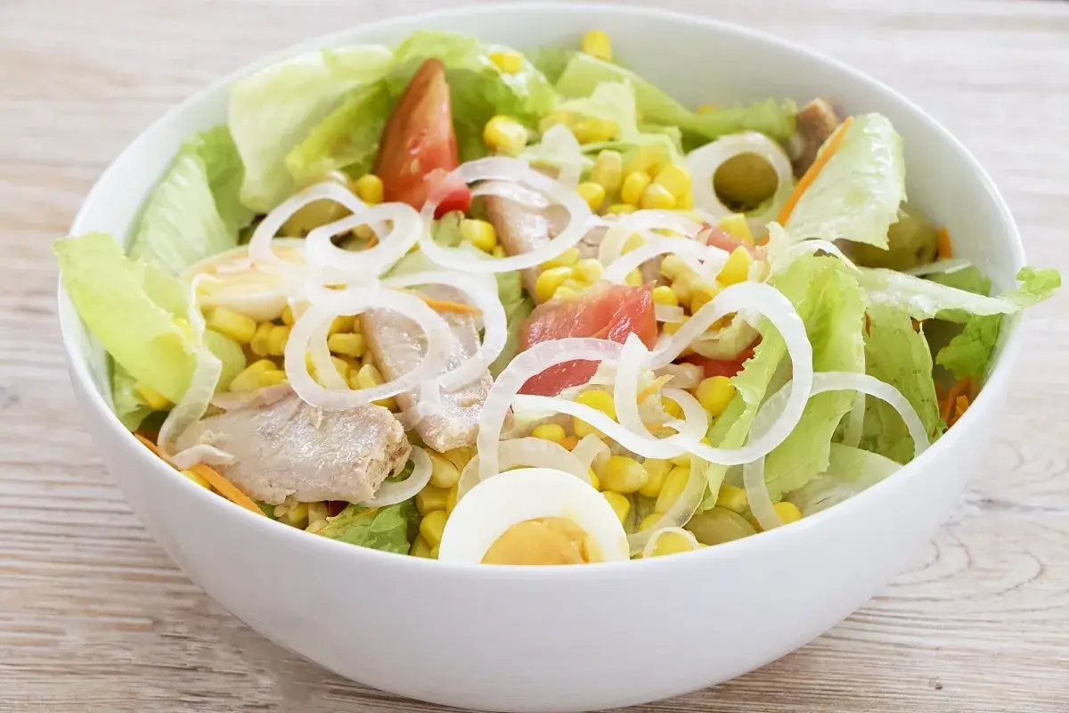 Deliciosa ensalada mixta con atún, huevo duro, maíz, tomate y cebolla, servida en un cuenco blanco sobre una mesa de madera.