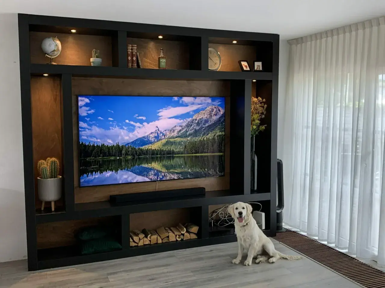 Ein Hund sitzt vor einer selbstgebauten TV-Wand mit vielen Fächern, die mit Deko, Büchern und Holz gefüllt sind.