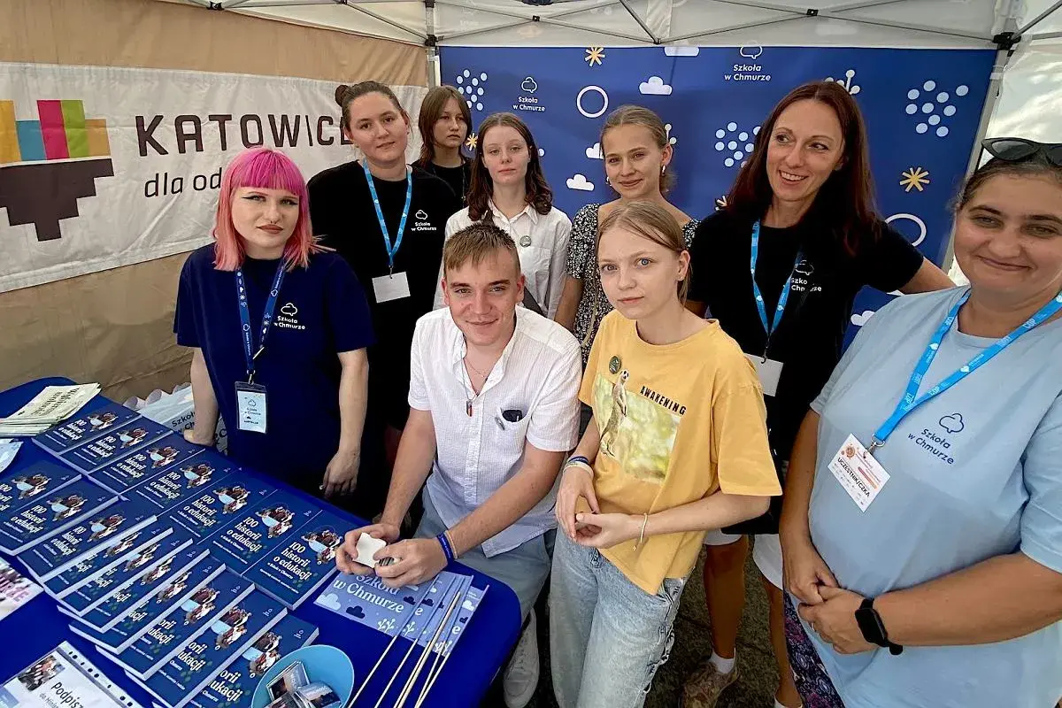 Grupa osób, w tym młodzież i dorosłych, stoi za stołem z książkami i materiałami promocyjnymi "Szkoła w Chmurze".