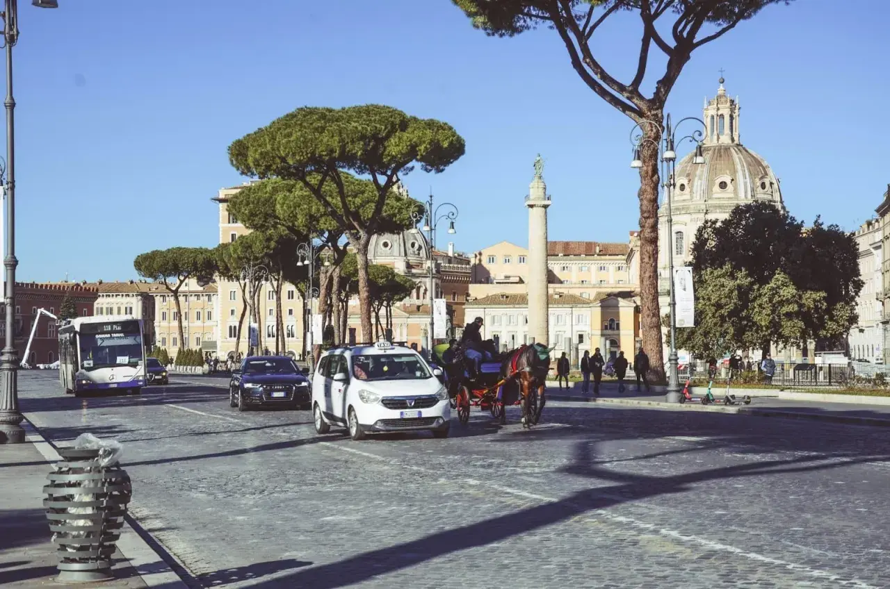 Un bus metro, taxi e carrozza a cavalli sfrecciano su un acciottolato a Roma, con edifici storici e pini marittimi sullo sfondo.