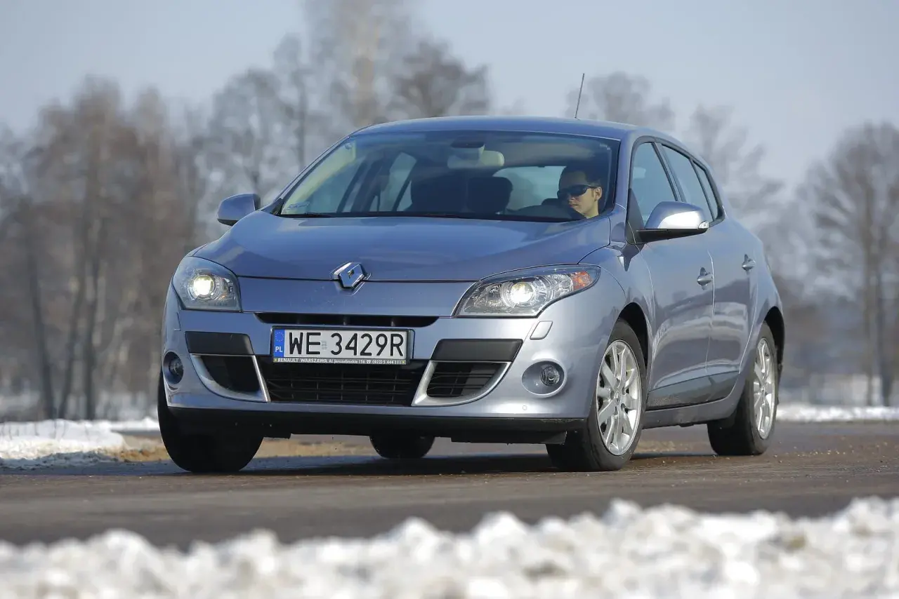 Srebrny Renault Megane 3 z włączonymi żarówkami przód, na drodze zimą.