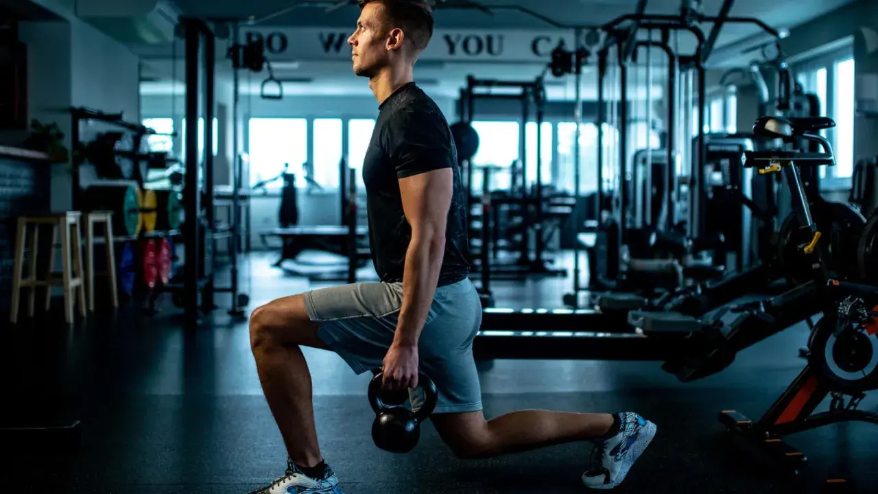 Mann macht Ausfallschritte mit Kettlebell im Fitnessstudio. Verschiedene Ausfallschritte Varianten trainieren Beine und Po.
