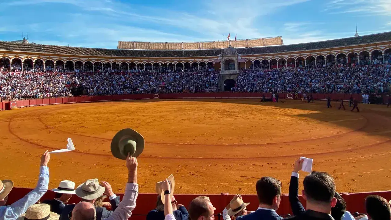 Espectadores en una **plaça de toros** levantan pañuelos y sombreros ante la arena dorada y el público en las gradas.