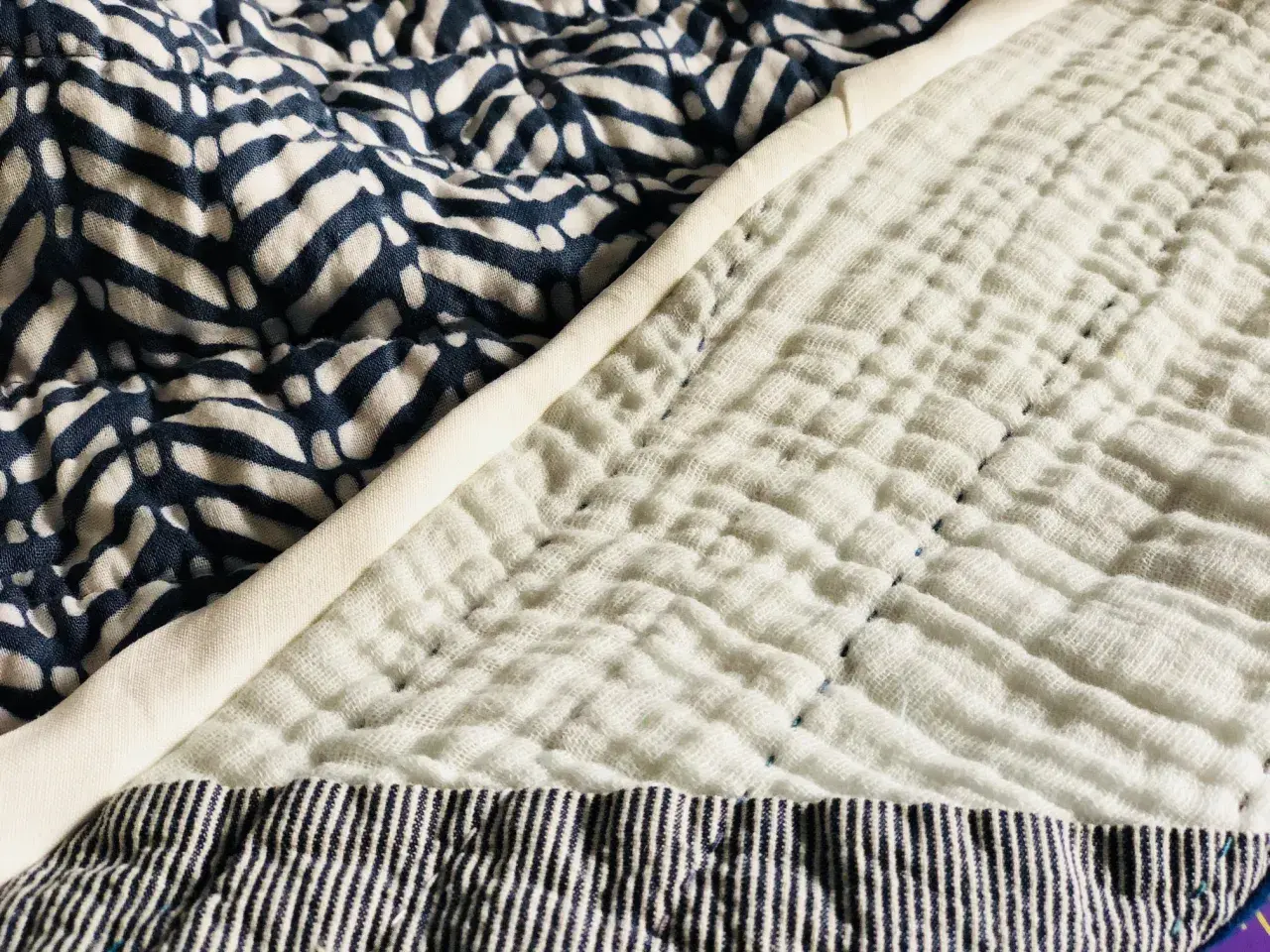 A close-up of a quilt shows a patterned blue fabric next to a textured white double gauze fabric, stitched with blue thread.