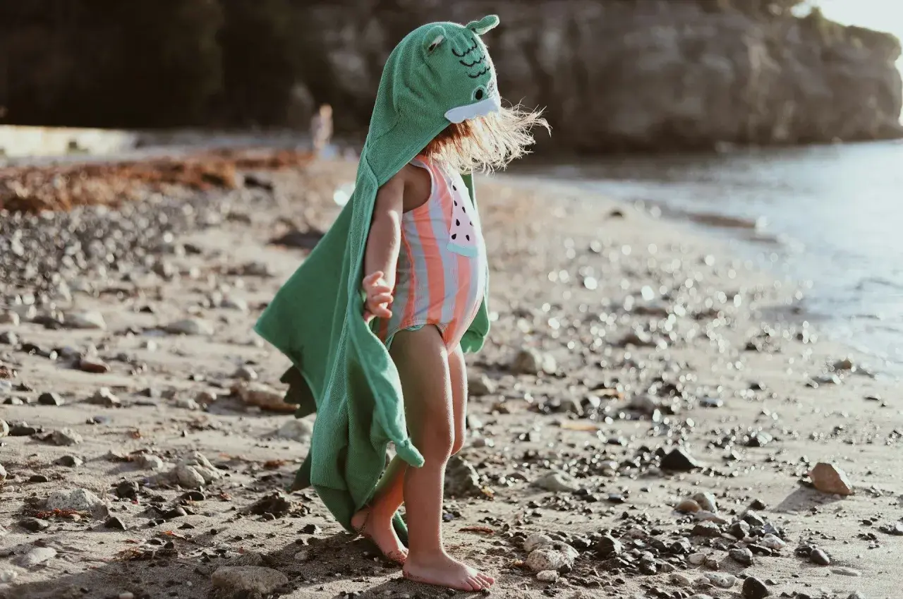 Liten badcape baby med dinosauriehuvud på stranden.