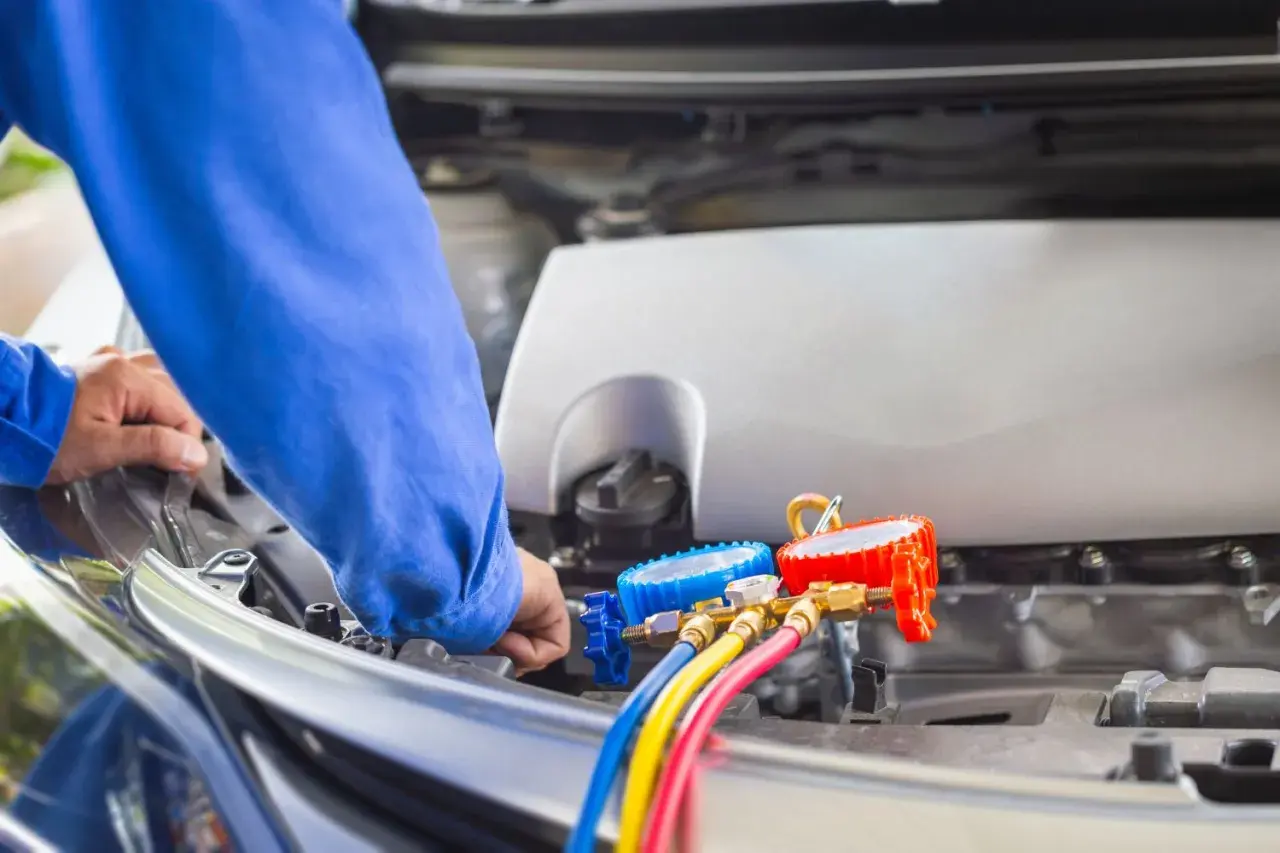 Mechanik w niebieskim kombinezonie wykonuje samodzielne nabijanie klimatyzacji w samochodzie, używając manometrów i kolorowych węży.