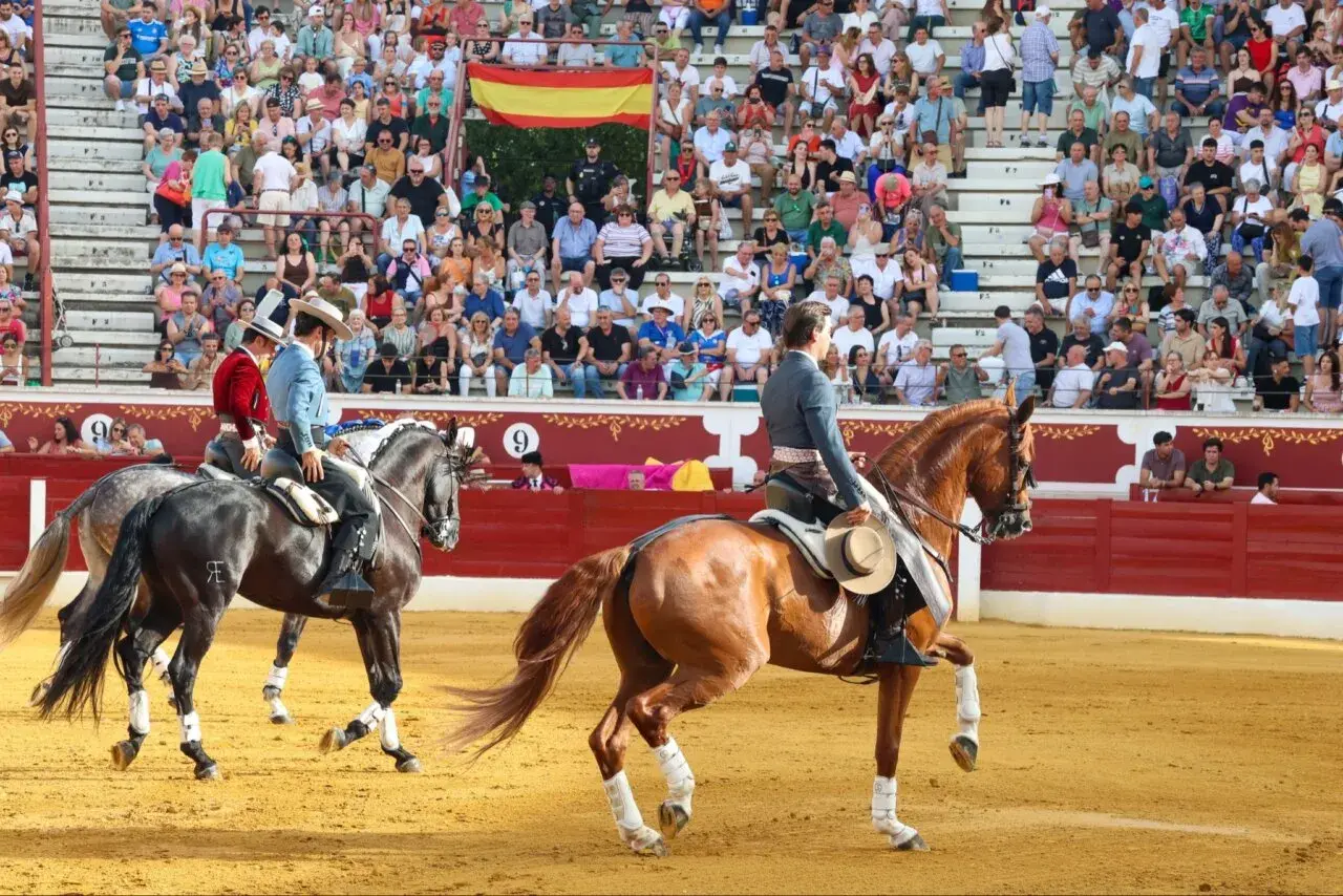 Dos jinetes, uno con chaquetilla roja y otro con azul, montan caballos en una plaza de toros. Bernardino Piriz, con sombrero, observa la escena.