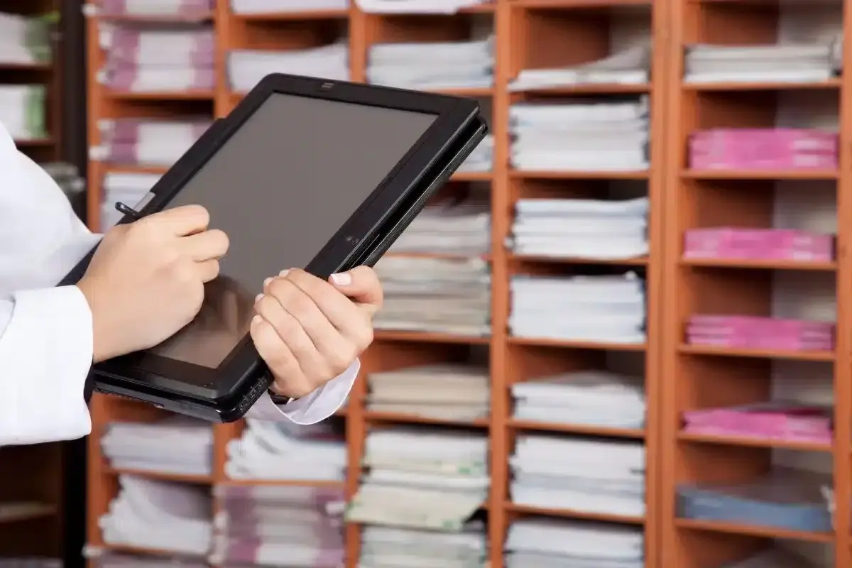Une personne utilise une tablette pour gérer des documents, illustrant l'archivage électronique, une obligation légale. Les étagères remplies de dossiers rappellent l'importance de la numérisation.