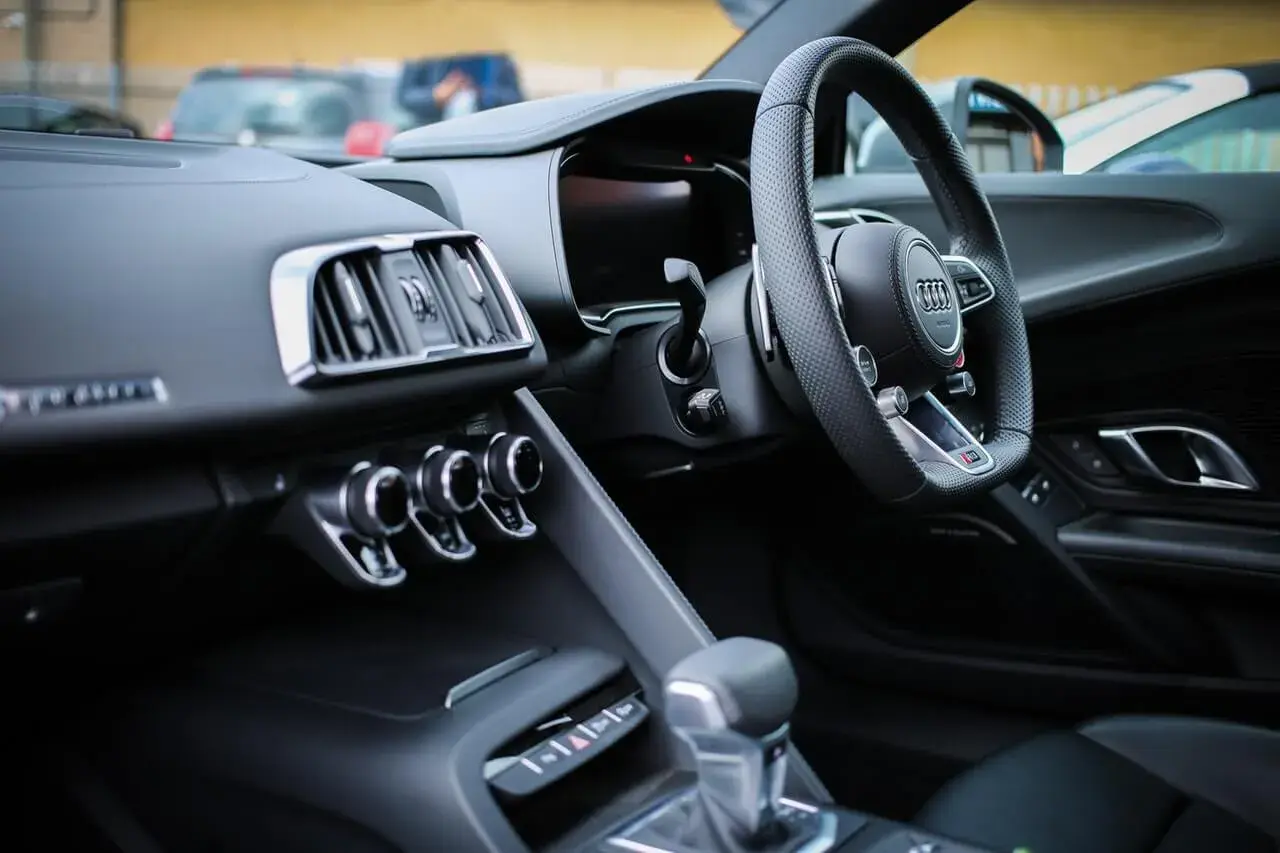 Interno di un'auto sportiva con volante a sinistra. Il cruscotto è scuro, con bocchette d'aria cromate e un display digitale. La leva del cambio è in primo piano. Perfetta per esplorare paesi con guida a sinistra.