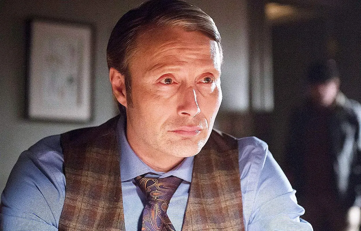 Mads Mikkelsen i en Mads Mikkelsen serie, klädd i skjorta, väst och slips. En annan person syns suddigt i bakgrunden.
