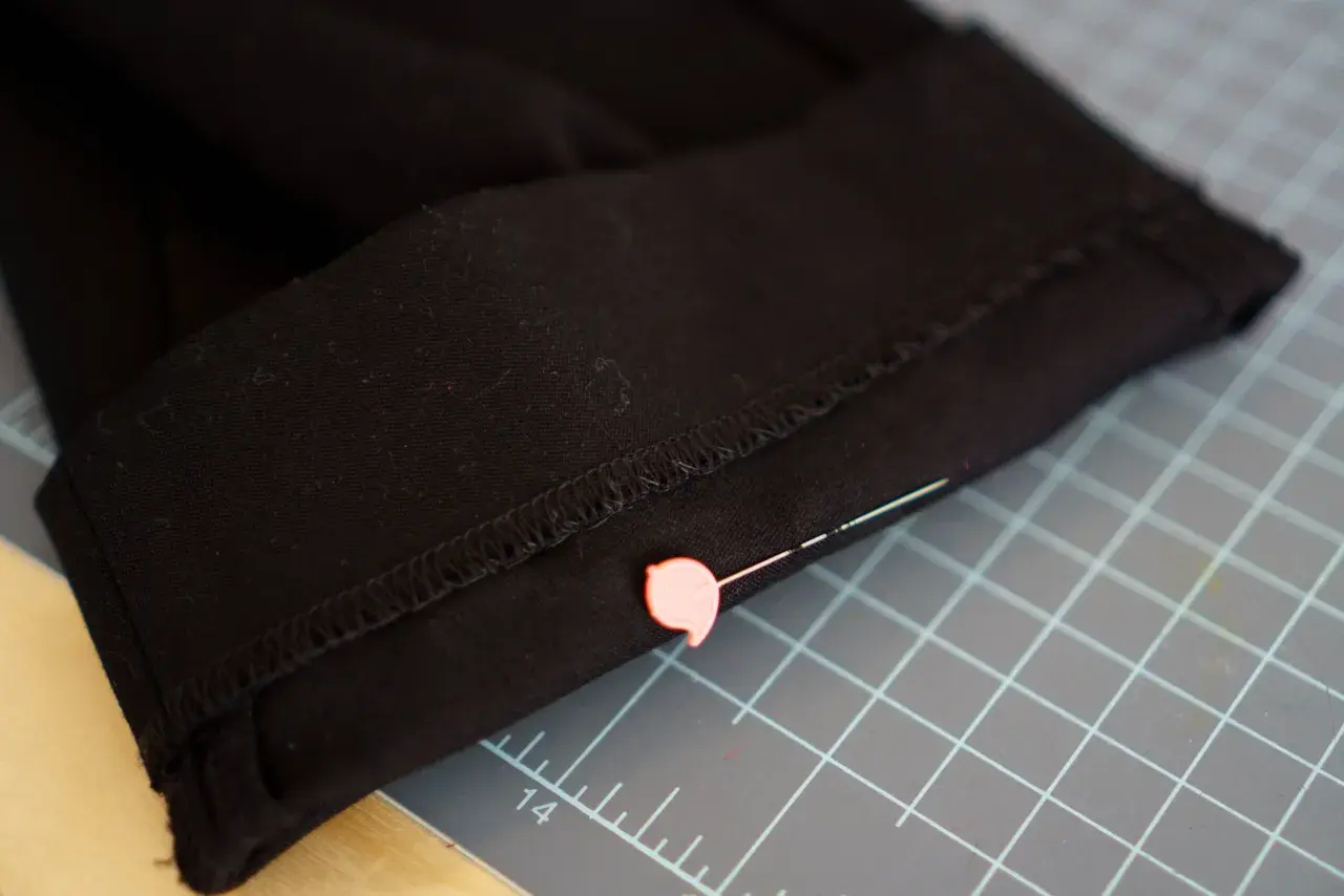 Close-up of black pants hem, pinned and ready for an invisible stitch. A pink pin secures the fabric on a cutting mat.