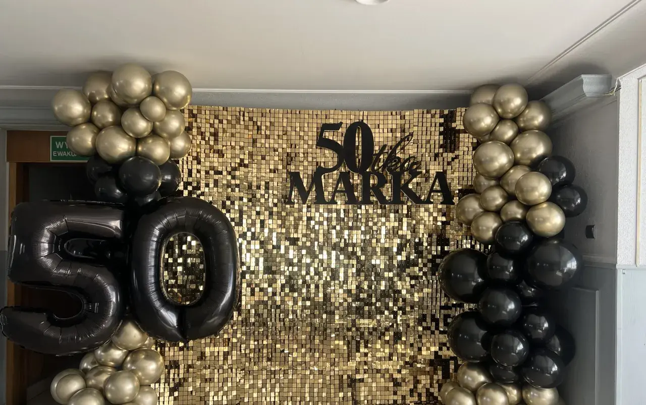 Złote i czarne balony otaczają błyszczące tło z napisem "50 lat Marka".