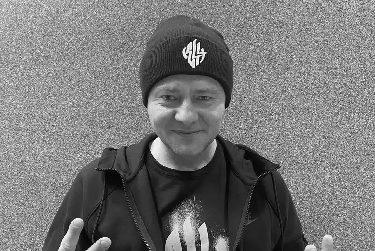 Mężczyzna w czapce z logo, w bluzie i koszulce. W tle szara ściana. Nie wiadomo, **joka na co zmarł**.