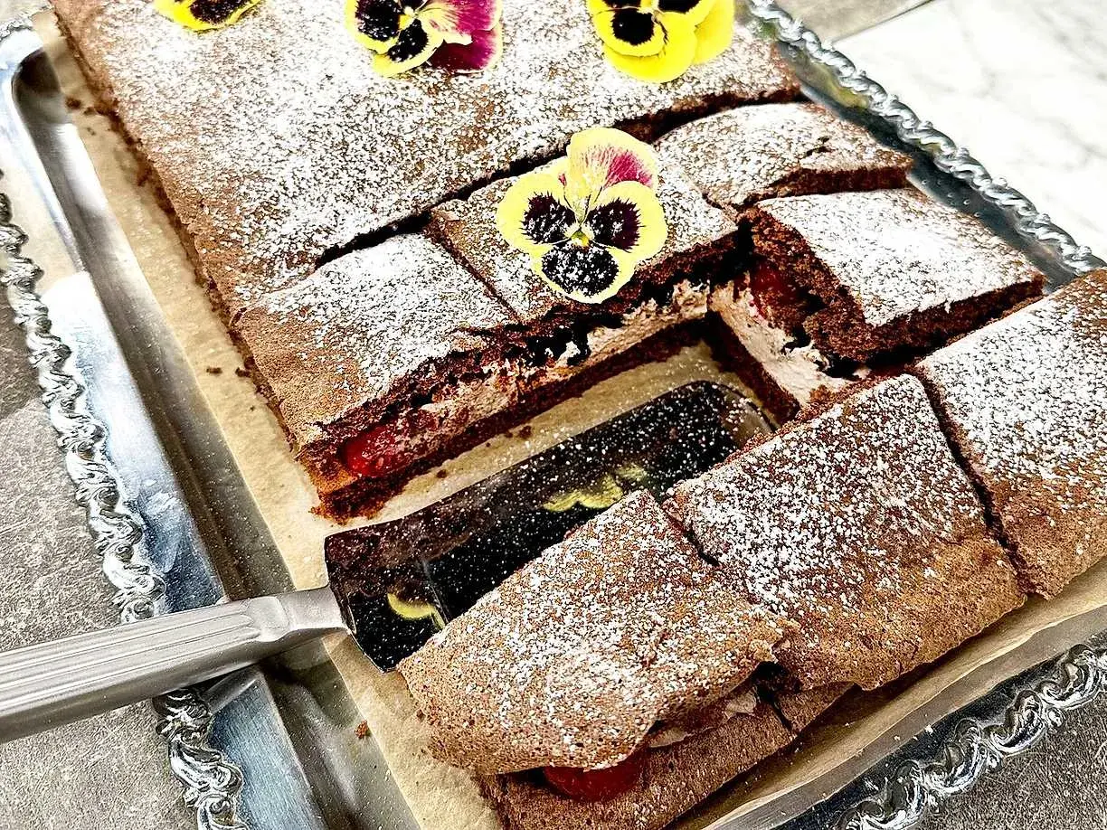 En chokladgrädde tårta skuren i bitar, pudrad med florsocker och dekorerad med violer. En tårtspade lyfter upp en bit.