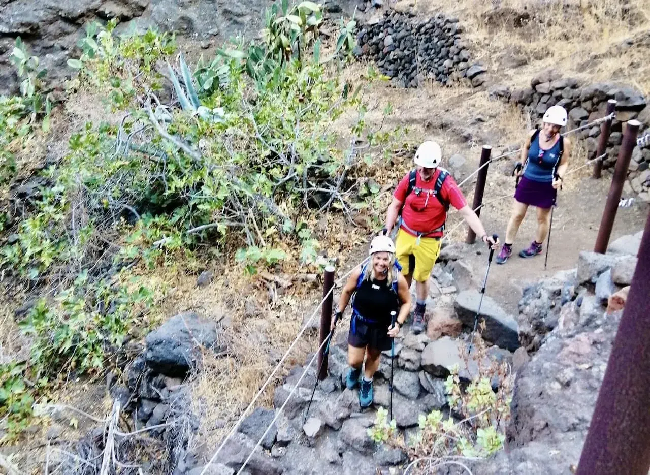 Wanderer steigen die felsige Masca Schlucht hinauf. Sie tragen Helme und Wanderstöcke.