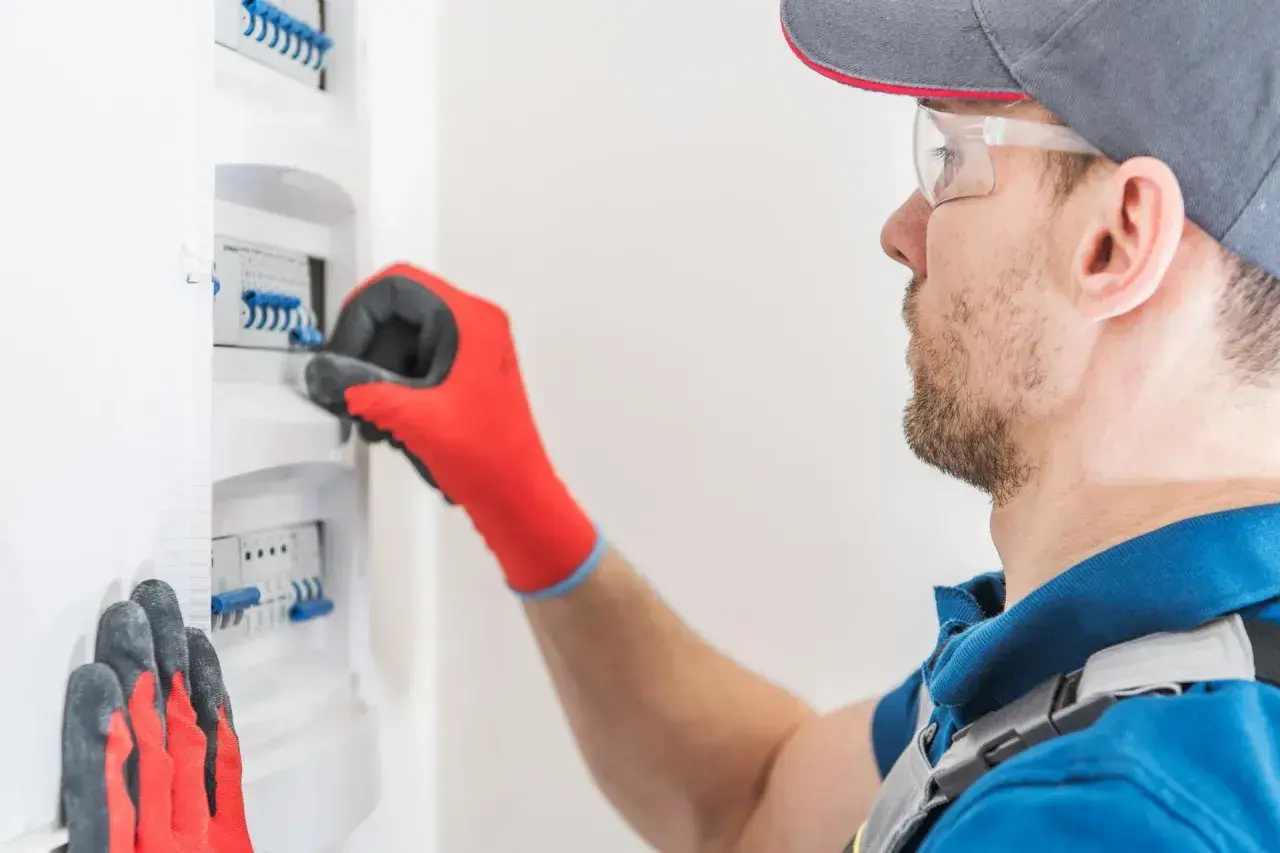 Elektryk w czerwonych rękawicach przeprowadza przegląd instalacji elektrycznej domu, sprawdzając bezpieczniki w skrzynce.