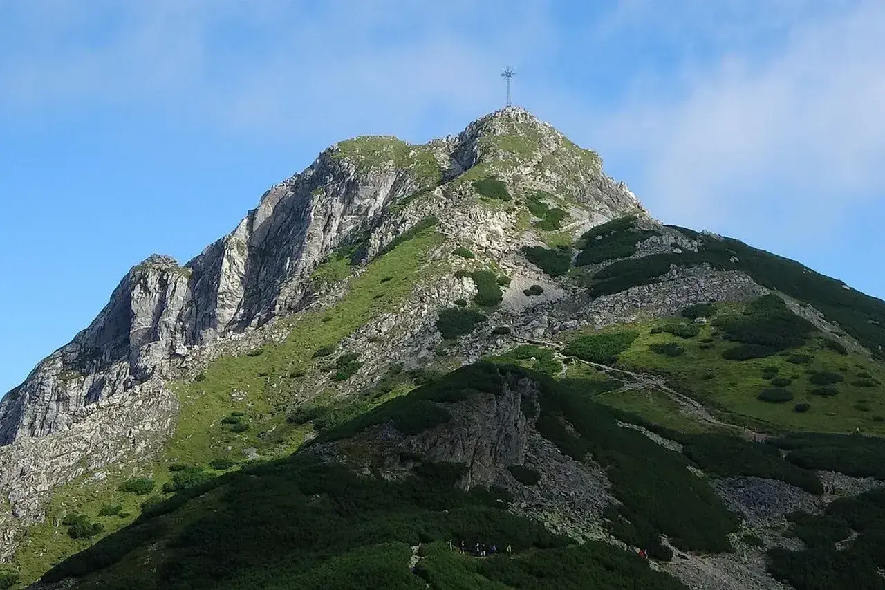 Widok na szczyt Giewontu z krzyżem, zielone zbocza i kamieniste ścieżki. W dole grupa turystów rozpoczyna wejście na Giewont.