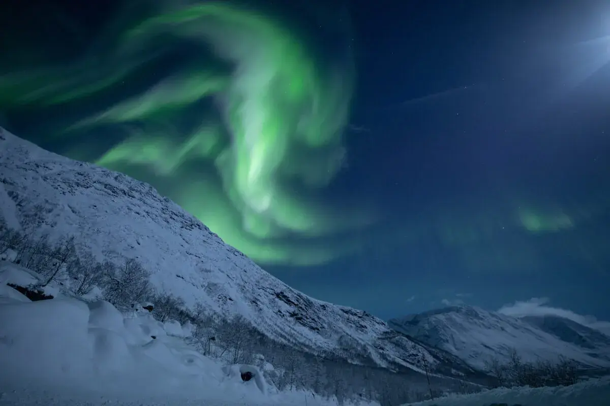 Grüne Nordlichter tanzen über schneebedeckte Berge bei Tromsø. Ein magischer Moment, der zu den Tromsø Geheimtipps gehört.