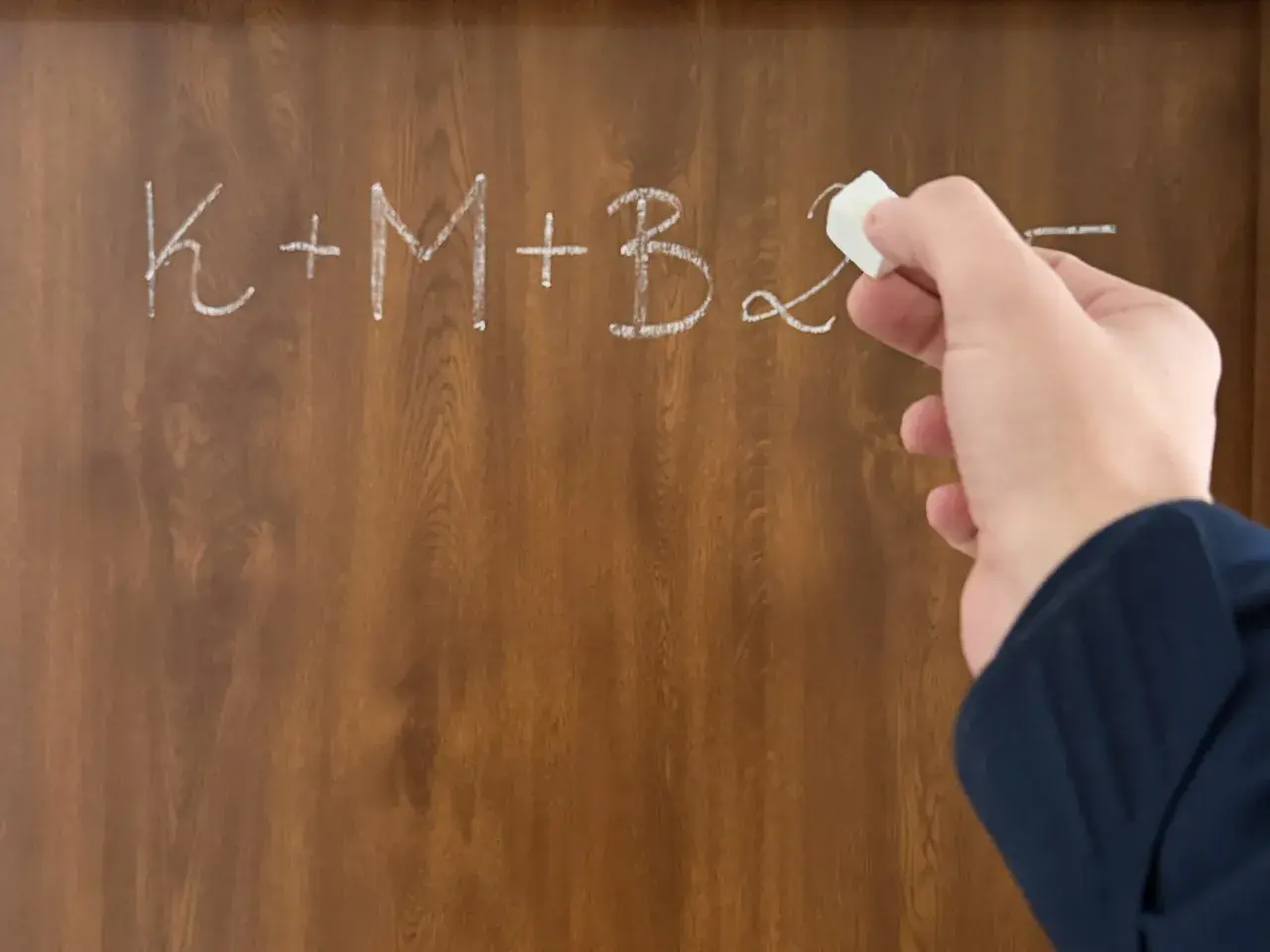 Dłoń pisze kredą na tablicy: K+M+B. Nagle przychodzi epifania, rozwiązanie problemu.