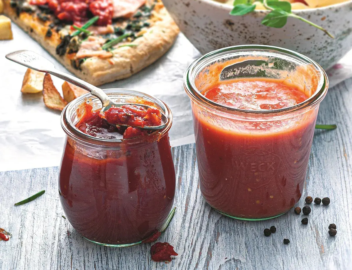 Dwa słoiki domowego sosu do pizzy, jeden z łyżką pełną aromatycznej masy. Obok kawałek pizzy i przyprawy.