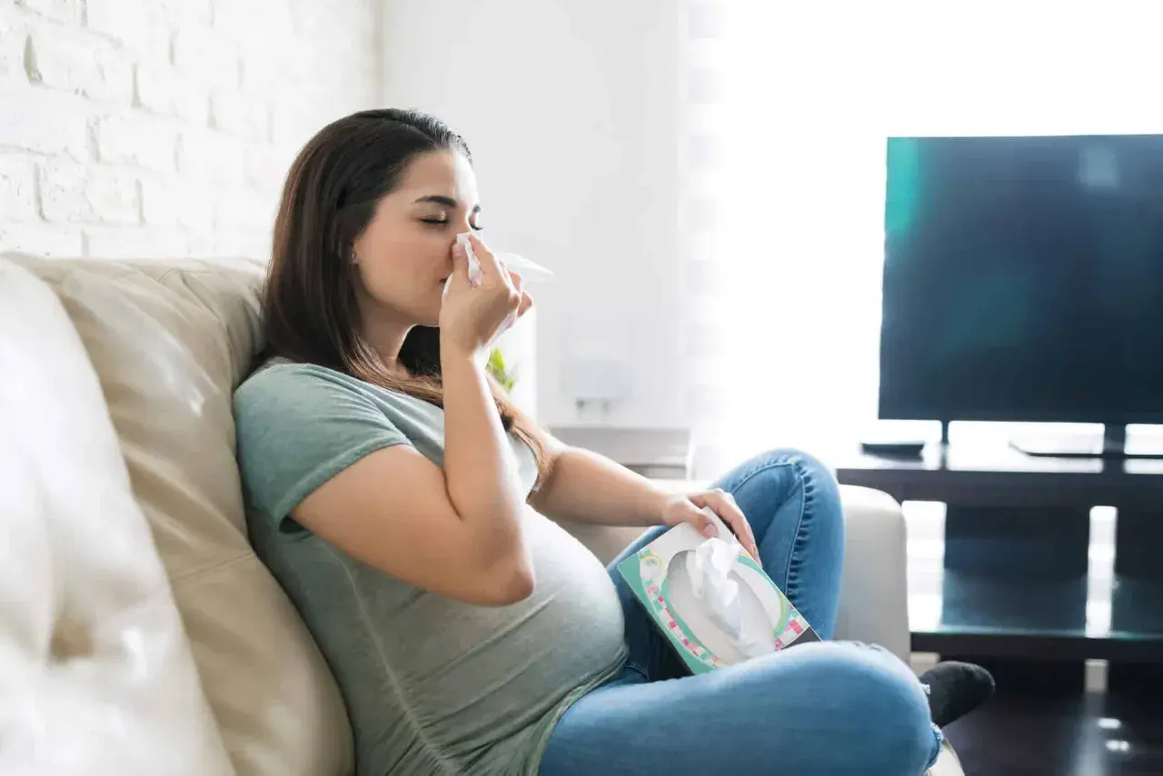 Femme enceinte s'essuyant le nez avec un mouchoir, inquiète d'un rhume enceinte danger pour bébé. Elle est assise sur un canapé, une boîte de mouchoirs à portée de main.