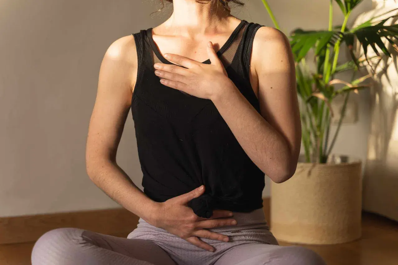 Une femme assise en tailleur, mains sur le ventre et la poitrine, pratique la respiration profonde. Une plante verte est visible en arrière-plan.