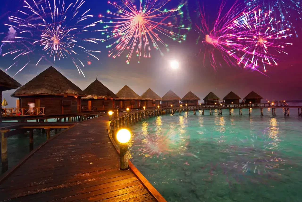 Bungalows auf Stelzen über türkisfarbenem Wasser, beleuchteter Steg, Feuerwerk am Nachthimmel. Hier ist es an Silvester warm und feierlich.