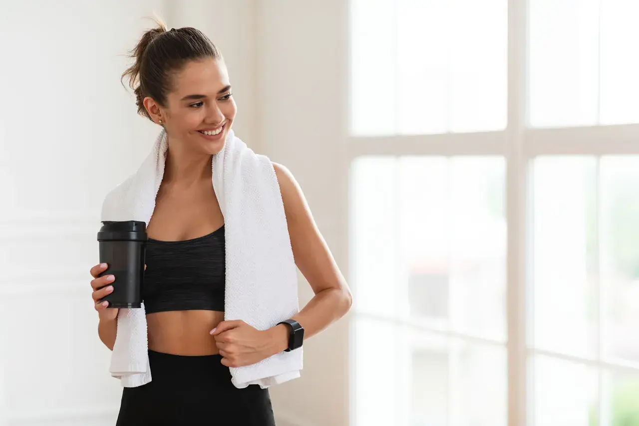 Lächelnde Frau mit Handtuch und Shaker nach dem Training. Sie fragt sich, wie viel Kreatin pro Tag für optimale Ergebnisse.