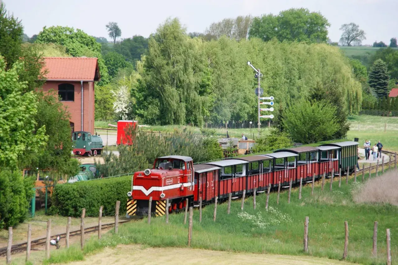 Czerwony pociąg z wagonami turystycznymi jedzie przez zielone tereny. W tle budynek i drzewa. To świetne pałuki atrakcje dla całej rodziny.