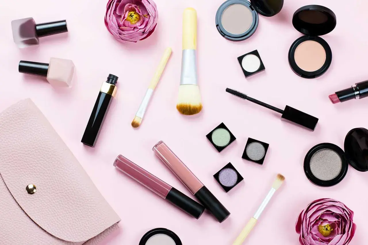 En rosa bakgrund med olika sminkprodukter som läppglans, mascara, puder och borstar. Perfekt smink i handbagage för en snabb touch-up.