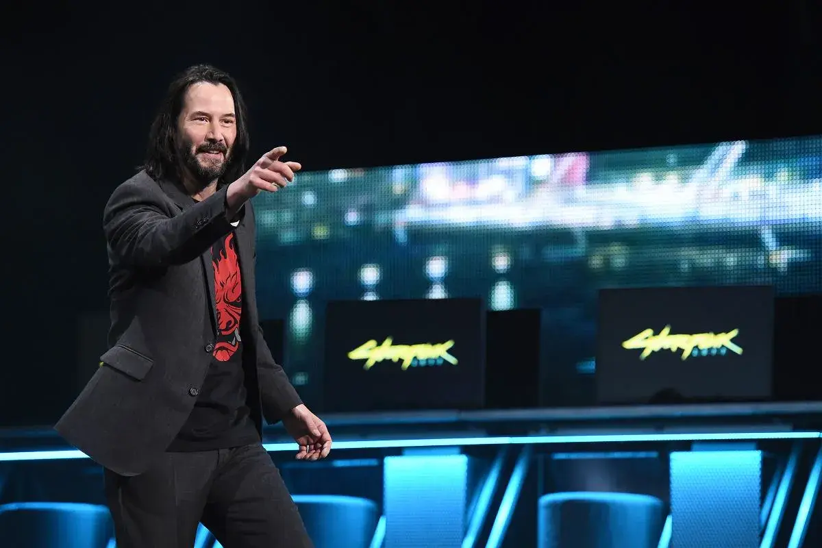 Keanu Reeves na scenie, wskazując palcem, w koszulce z logo Cyberpunk 2077. Gra zarobiła miliony, a on jest jej twarzą.