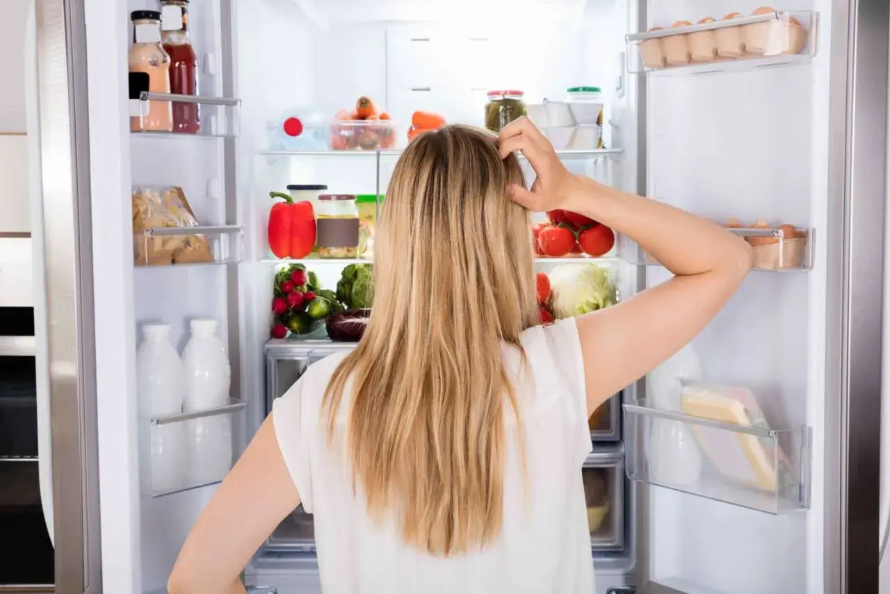 Donna confusa davanti al frigo pieno di cibo, con il desiderio di mangiare e non ingrassare, ma lo stress la blocca.