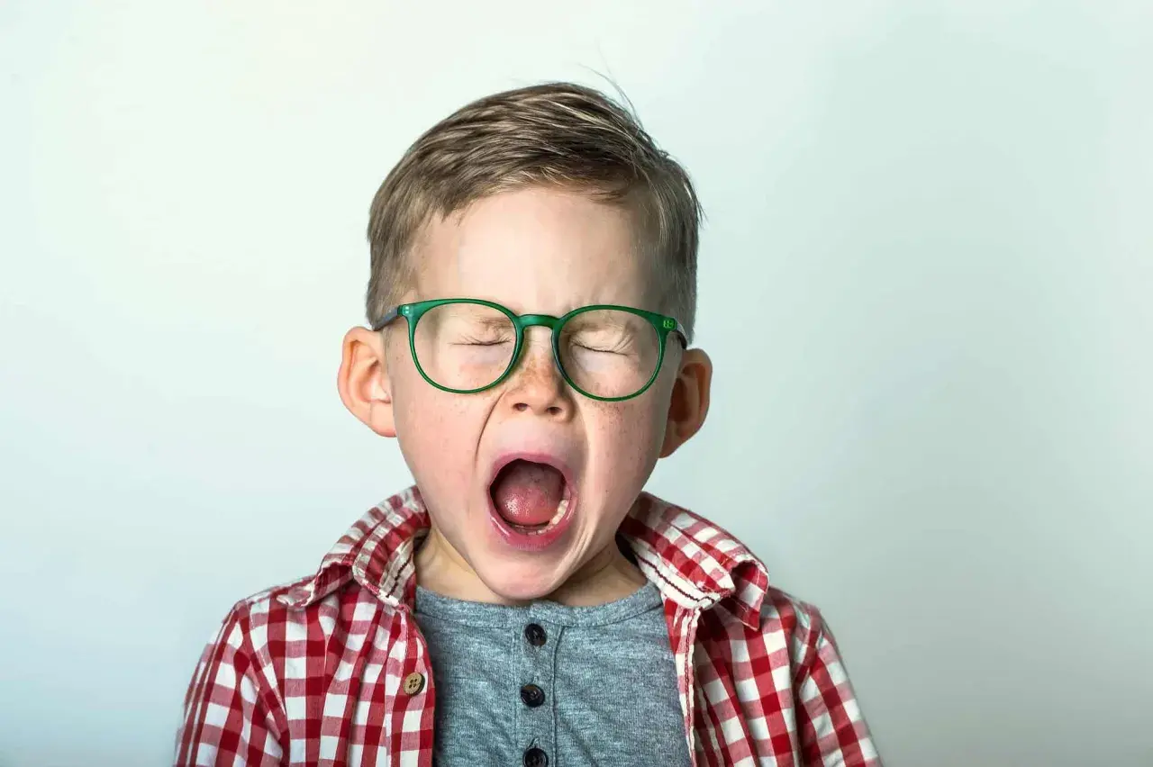 Niño con gafas verdes y camisa a cuadros, con los ojos cerrados y la boca abierta, expresando una emoción intensa, como podría ocurrir en el síndrome de Tourette en niños.