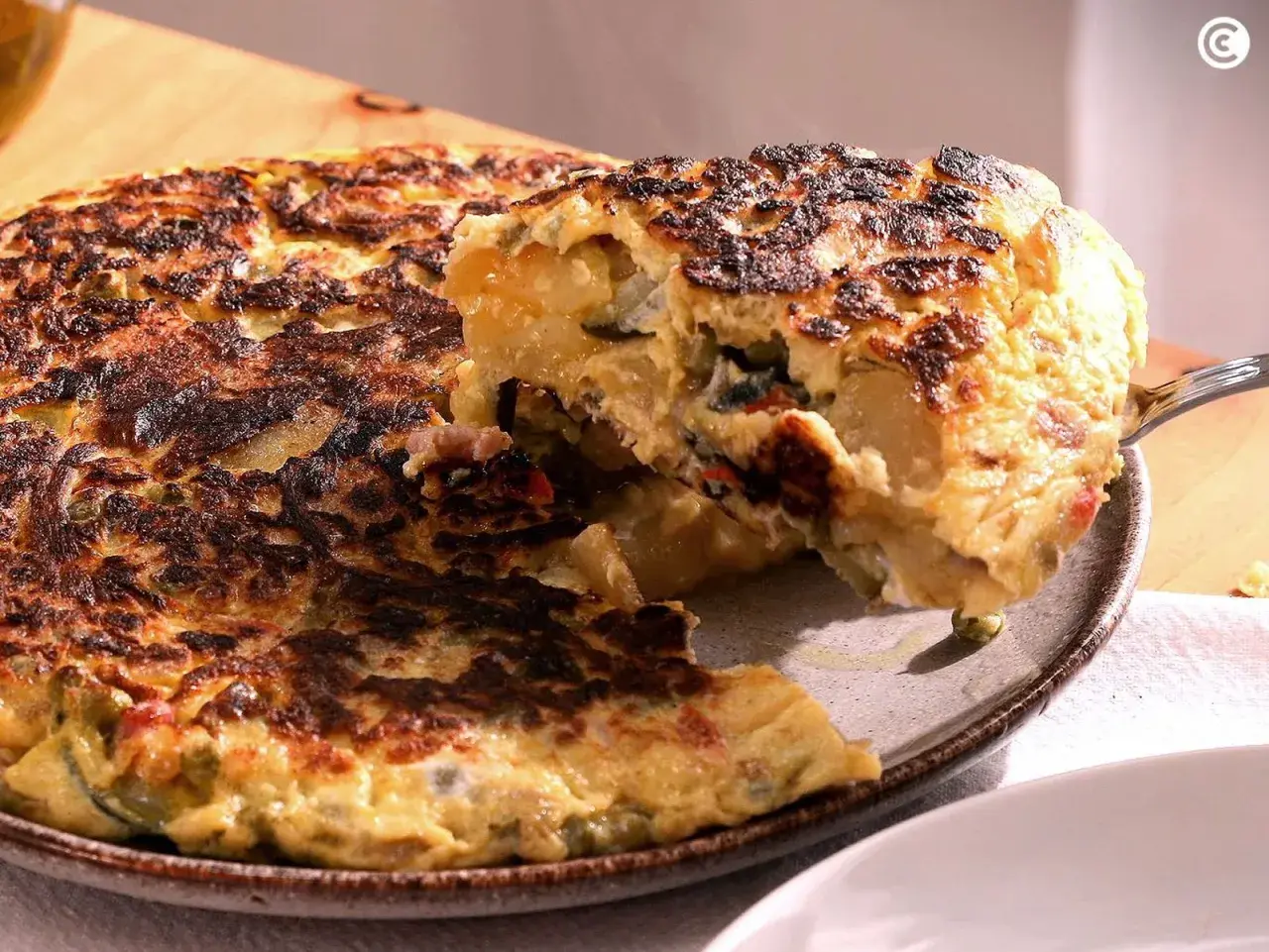 Una porción de tortilla paisana, dorada y jugosa, se levanta con un tenedor, revelando patatas, pimientos y huevo.