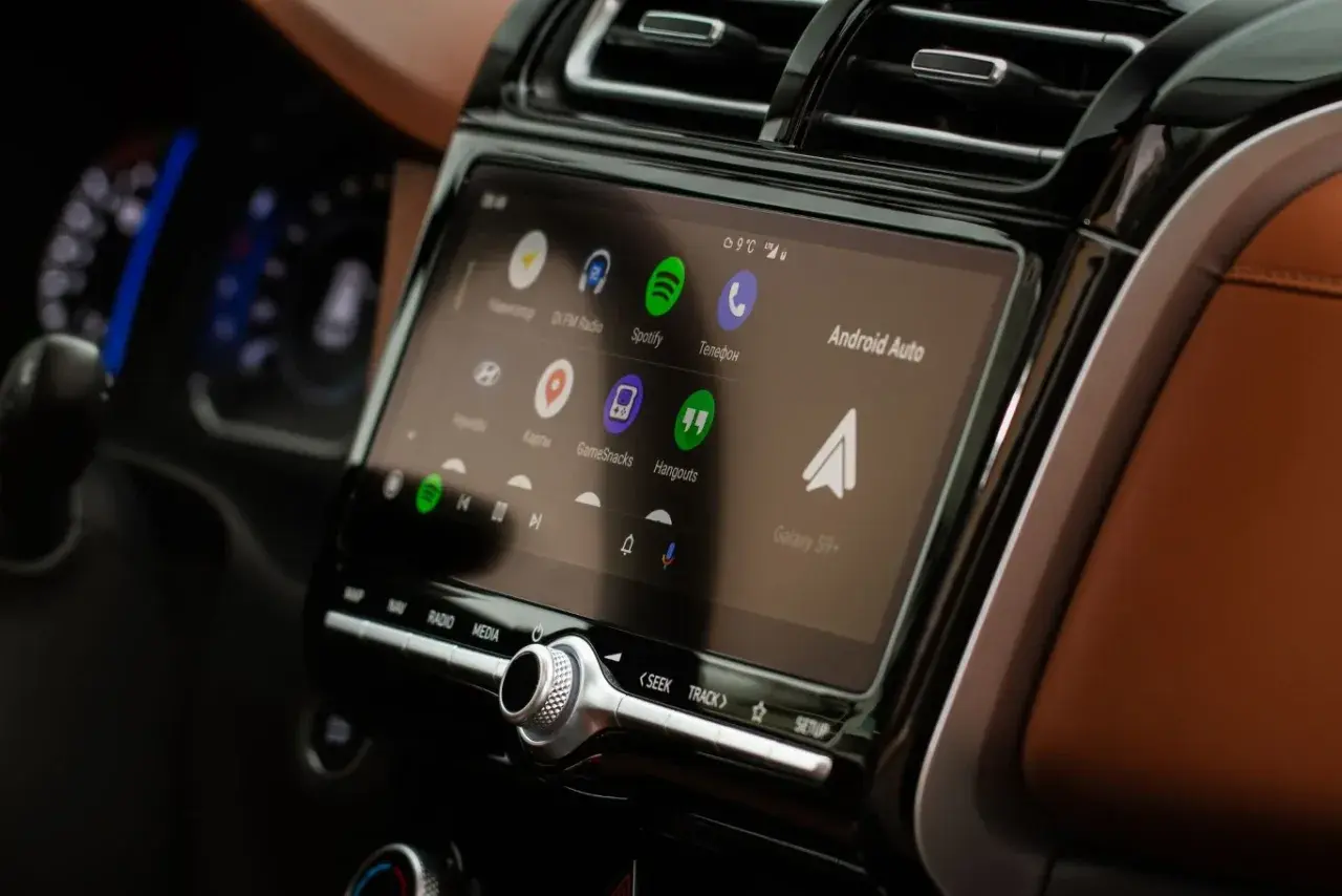 Ekran multimedialny z Android Auto, pokazujący ikony aplikacji: Spotify, Telefon, Mapy. W tle widać fragment deski rozdzielczej.