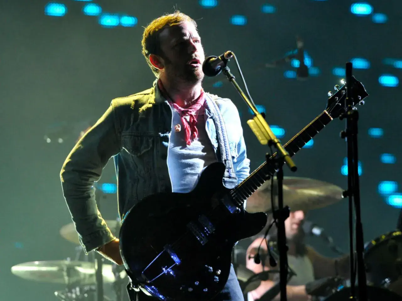 Wokalista Kings of Leon Polska na koncercie, gra na gitarze elektrycznej, w dżinsowej kurtce i czerwonej chuście.