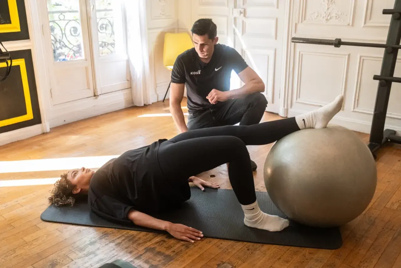 Un homme observe une femme faire un pont sur un tapis, les pieds sur un ballon de gym. Un exercice pour le bienfait d'étirement et de renforcement.