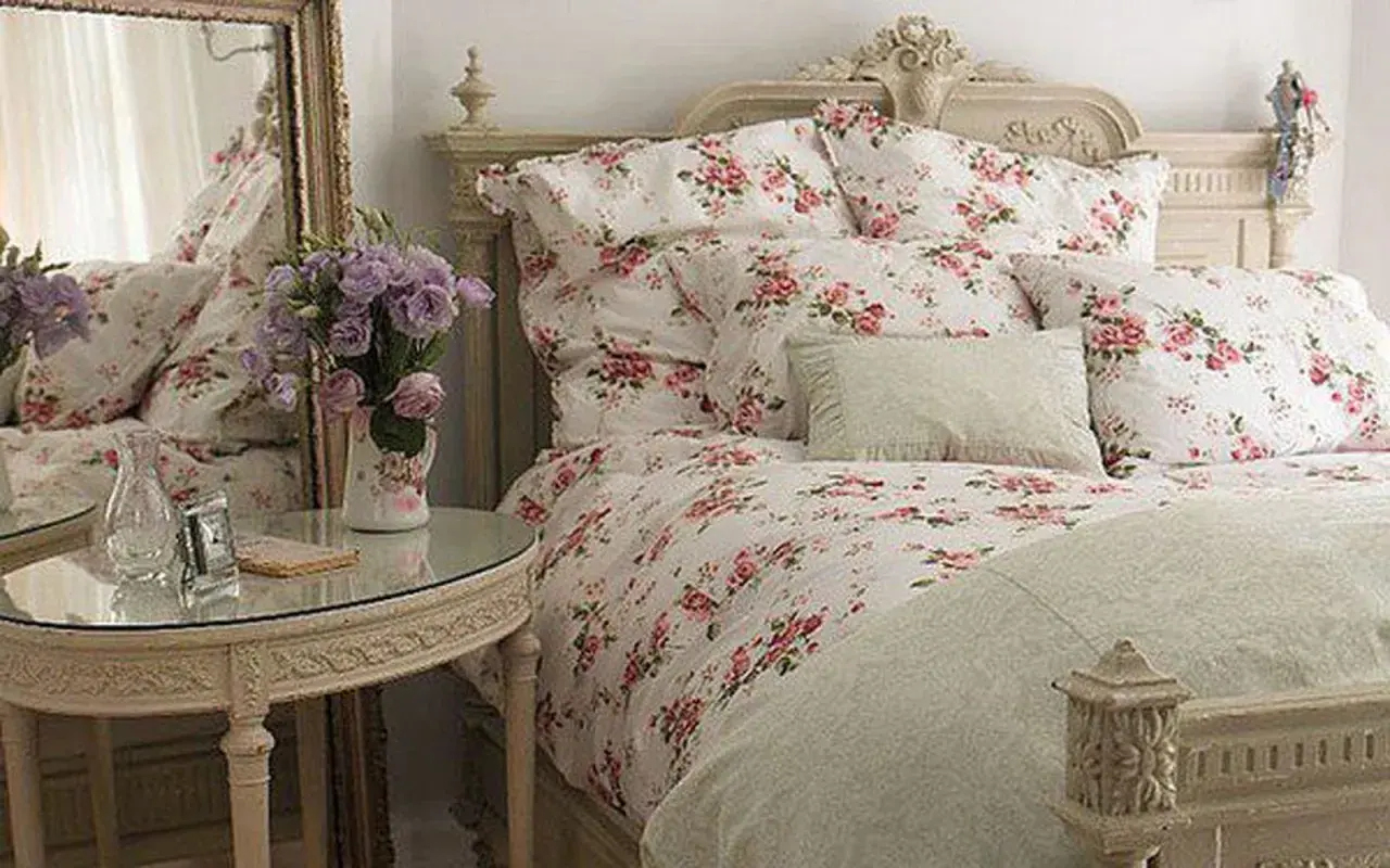 En säng med blommig shabby chic inredning, prydd med rosor och lavendel. Ett runt spegelbord med en vas full av blommor.