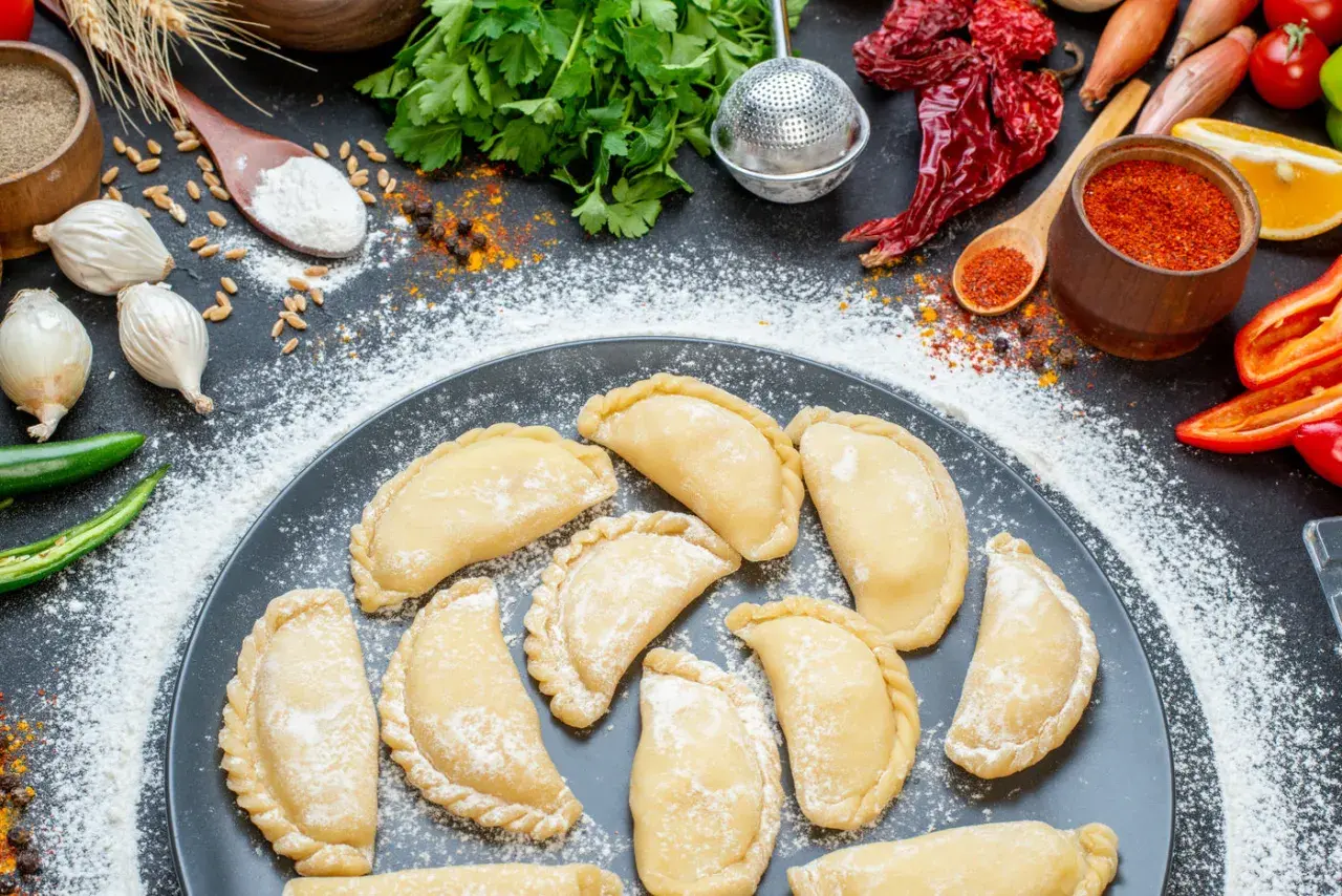 Gotowi, by nauczyć się, jak zrobić pierogi? Na talerzu surowe pierożki, obok mąka, cebula, papryczki chili i przyprawy.