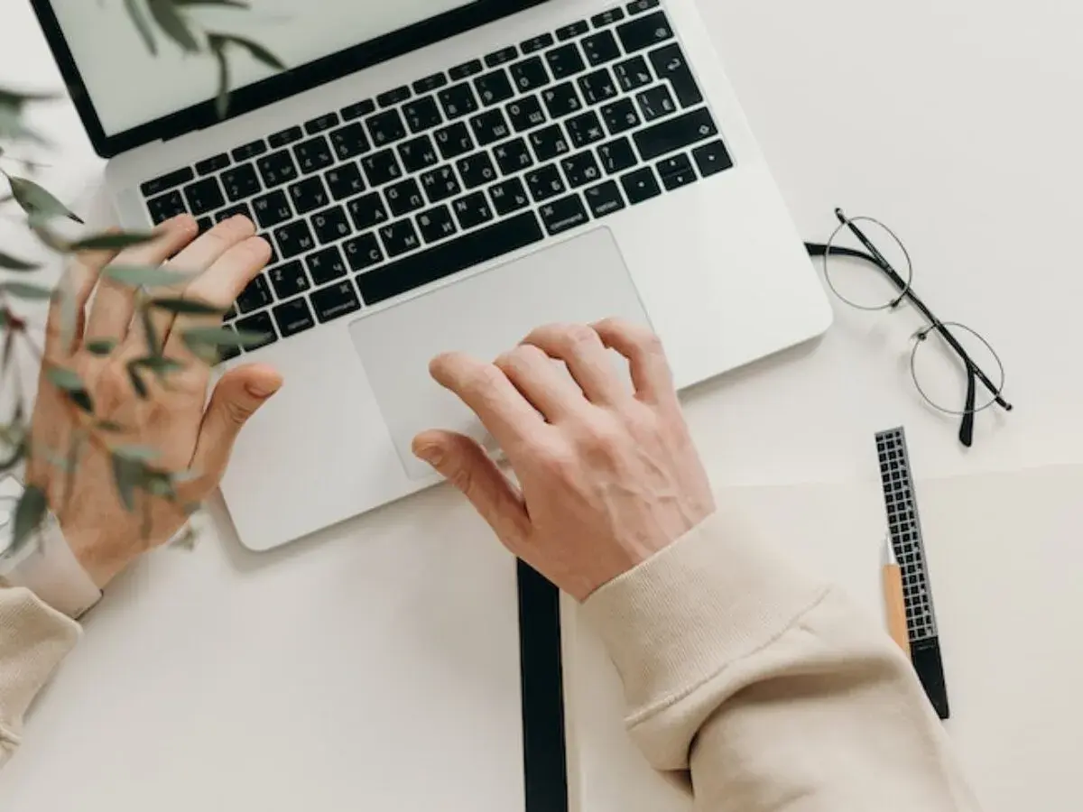 Ręce piszą na klawiaturze, obok leżą okulary i notatnik. Idealny tani laptop na studia, by zacząć naukę.