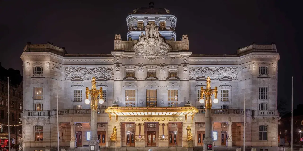 Kungliga Dramatiska Teatern i Stockholm, upplyst på natten. En storslagen byggnad med detaljerad fasad och gyllene utsmyckningar.